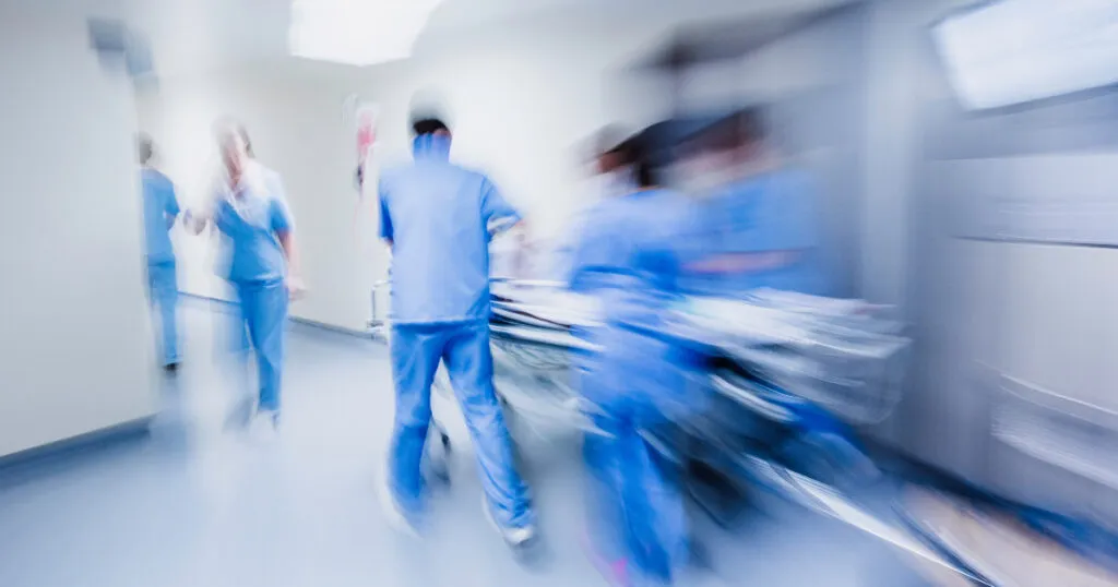 Motion-blurred image of hospital staff in blue scrubs rushing a patient on a gurney down a bright corridor.