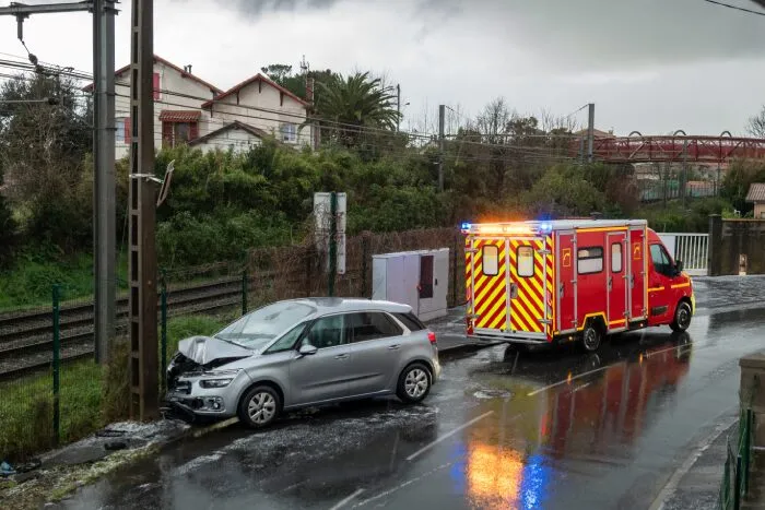 Silver car crashed into a pole near train tracks, with an emergency response vehicle parked nearby with flashing hazard lights.