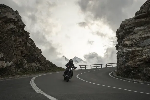 A motorcyclist wearing a helmet and dark gear rides along a winding, mountainous road with steep rock cliffs on both sides.