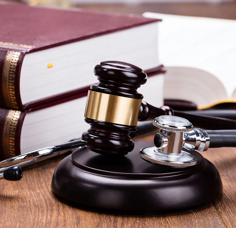 A gavel rests on a wooden block, positioned next to a stethoscope and two large, leather-bound books on a wooden desk.