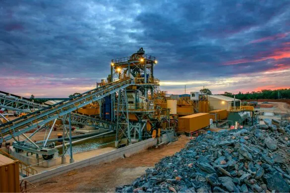 An industrial aggregate processing or mining facility with conveyors and crushing equipment is shown at dusk, with large piles of crushed rock in the foreground.