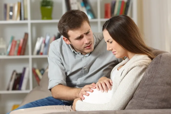 A concerned man comforting a pregnant woman who appears to be in pain or distress, sitting together on a couch.