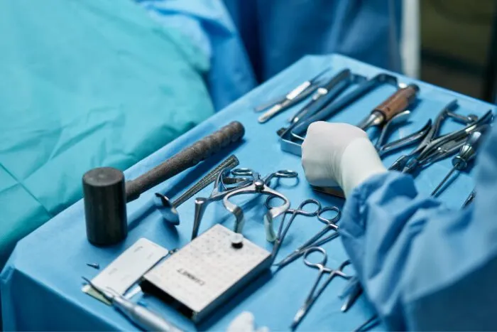 A medical person holding a variety of specialized surgical tools, including scissors, retractors, and a small mallet, likely for an orthopedic procedure.