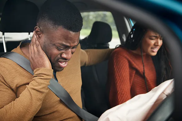 An image of a man and a woman sitting in a car, holding the back of their necks in pain.