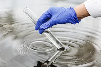 An image of a person wearing a lab gown and blue gloves, pouring water into a tube.