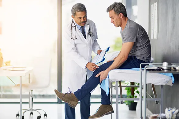 A male patient sits on an examination table, holding his injured leg, while a male doctor in a white coat examines the knee and holding patient document.