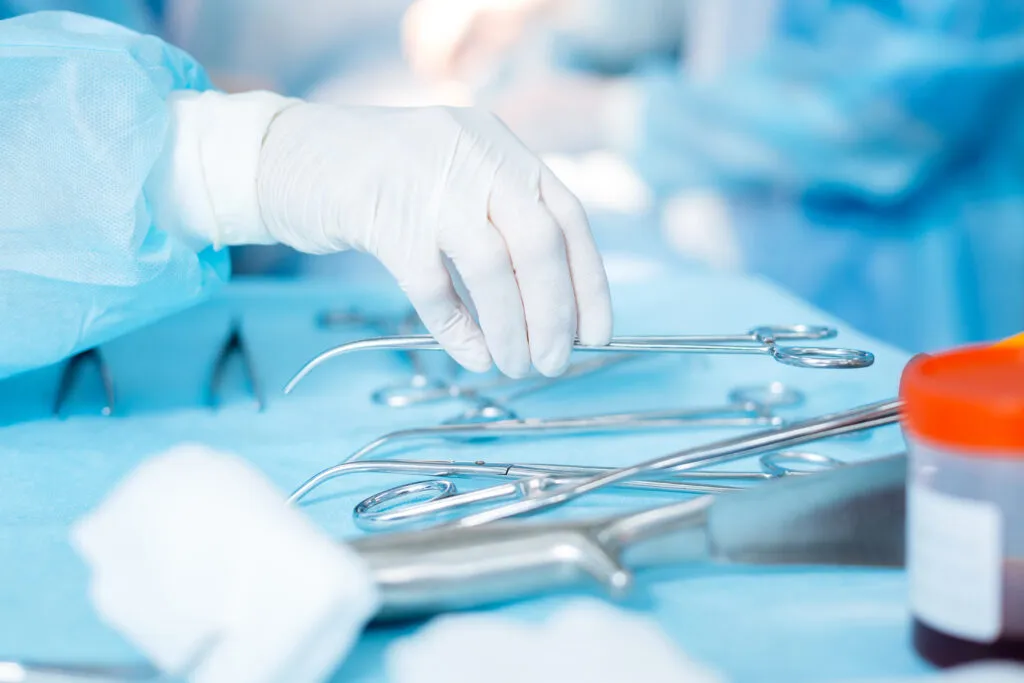 A medical professional, wearing white sterile gloves, reaching for a metal surgical instrument, such as forceps or scissors, laid out on a sterile blue cloth during an operation.